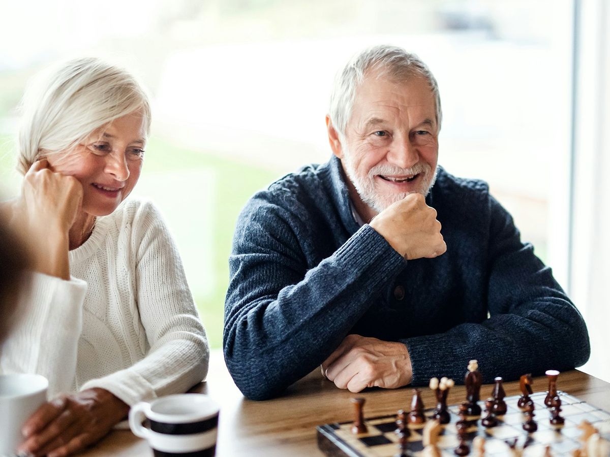 Two older adults smile across a chessboard beside a bright window, mid-game with a friend.